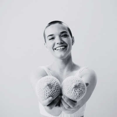 Smiling woman holding two knitted breast-shaped models toward the camera.